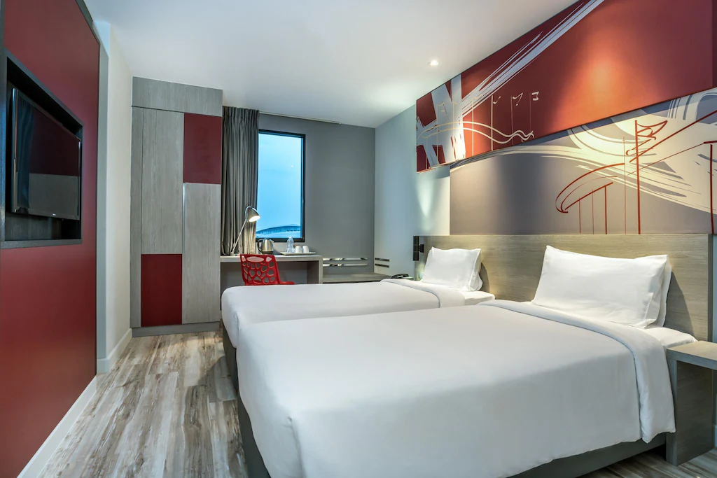 Modern hotel room with two single beds, white bedding, a wall-mounted TV, a desk with a lamp and chair by the window, and abstract red and gray artwork on the wall above the beds.