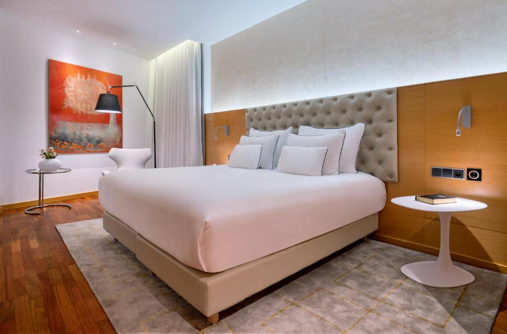 A modern hotel room with a large bed featuring white linens and a tufted headboard, flanked by two round white side tables. The room has wooden floors, a rug, modern art on the wall, and soft lighting.