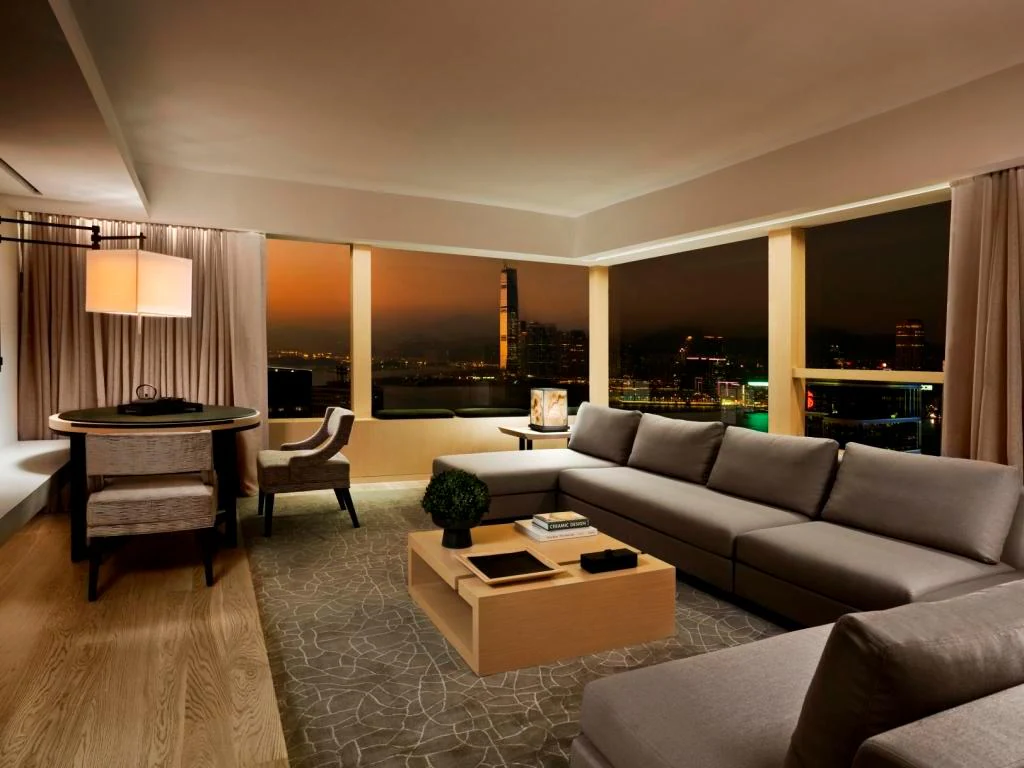 Modern living room with a large gray sectional sofa, wooden coffee table, patterned rug, dining area, and floor-to-ceiling windows showing a cityscape at dusk.