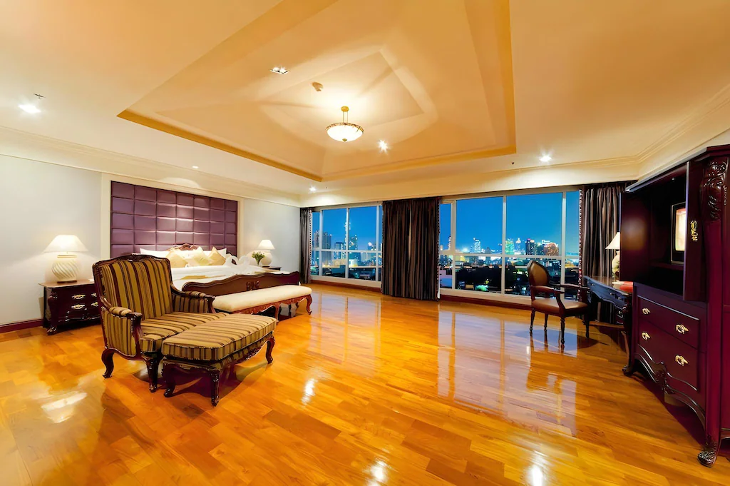 Spacious luxury bedroom with polished wooden floors, a large bed with a purple headboard, classic armchair with ottoman, desk by wide windows overlooking a city skyline at night, and elegant dark wood furniture.