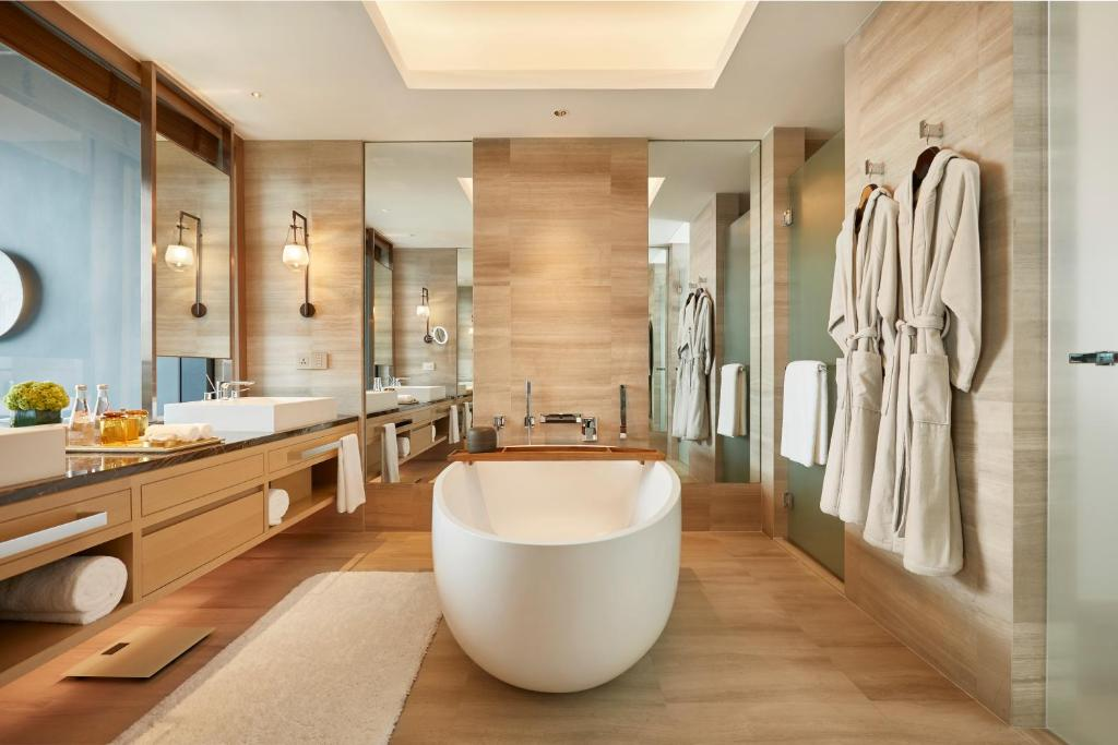 A modern, spacious bathroom with a freestanding white bathtub, double sinks, large mirrors, light wood floors and walls, glass shower, towel racks, and two hanging bathrobes. Soft lighting creates a relaxing atmosphere.