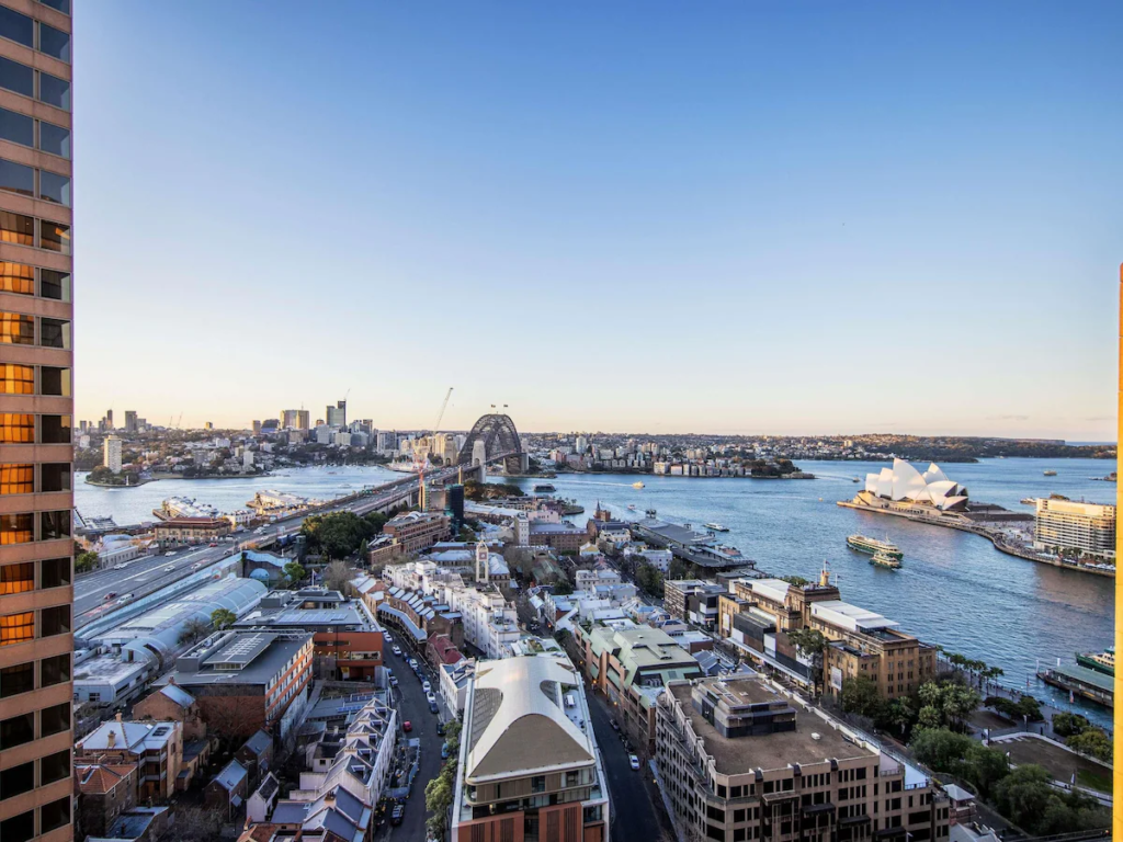 Aerial view of Sydney featuring the Sydney Harbour Bridge, the Opera House, and buildings along the waterfront under a clear blue sky. Ferries and boats are visible in the harbor.