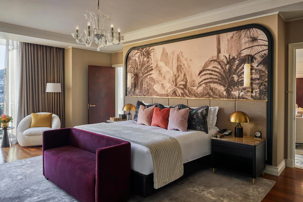A stylish hotel bedroom with a large bed, colorful pillows, a maroon bench, gold lamps, and a nature-themed mural above the headboard. The room has a chandelier, modern furniture, and large windows with light curtains.
