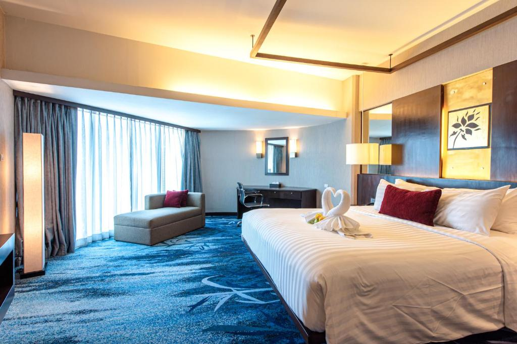 Modern hotel room with a large bed, blue carpet, a desk, and a lounge chair by floor-to-ceiling windows. Towels on the bed are folded into two swan shapes, and soft lighting creates a cozy atmosphere.