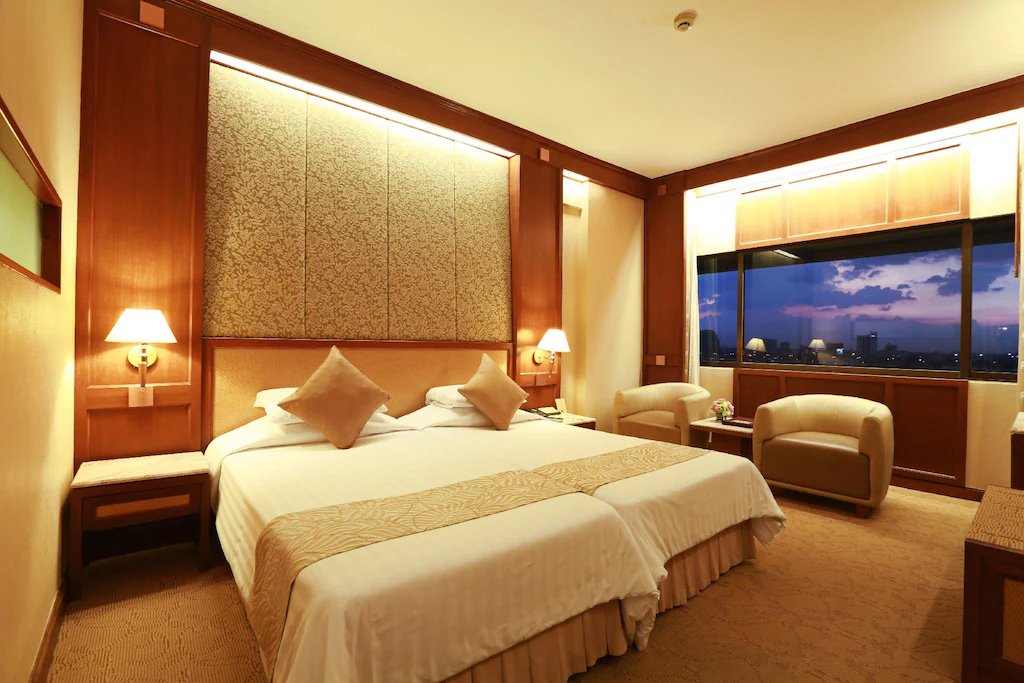 A hotel room with two twin beds pushed together, beige bedding, two armchairs by a large window showing a city skyline at sunset, warm lighting, and wooden furnishings.