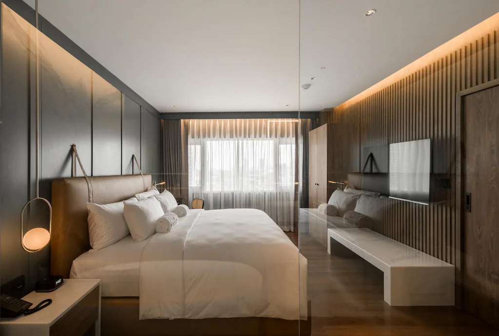 A modern hotel room with a large bed, white bedding, and rolled towels. The room features wood paneling, a wall-mounted TV, a white bench, and a large window with sheer curtains letting in natural light.