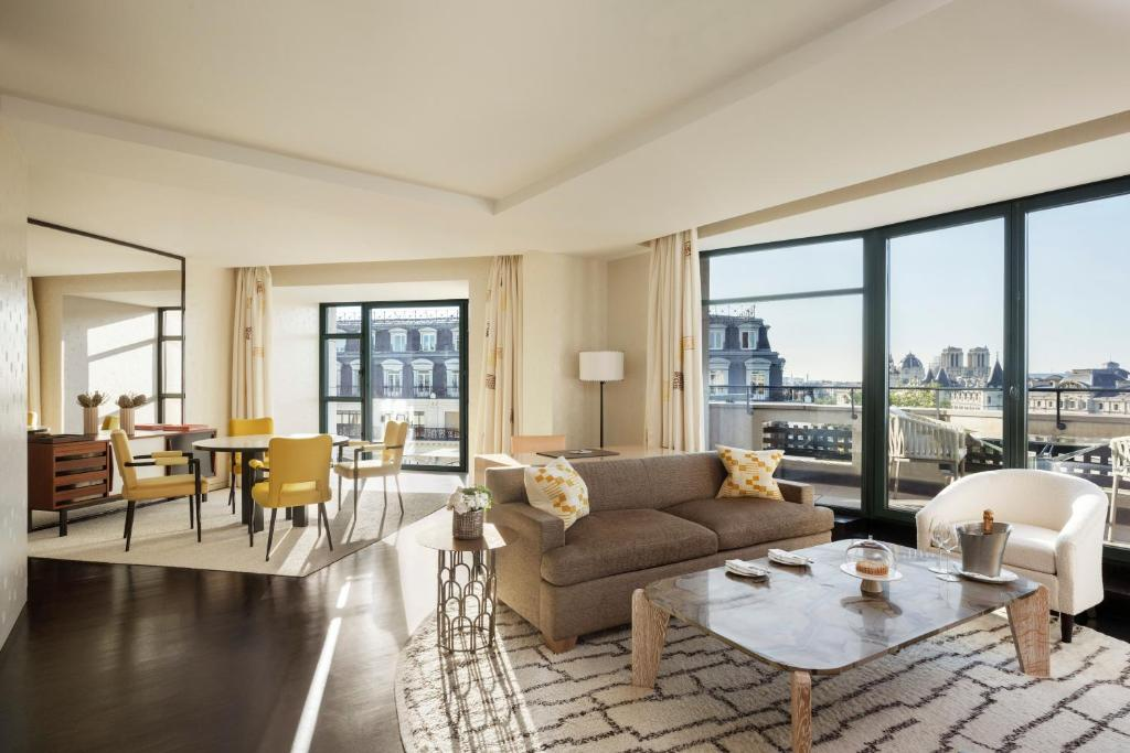 A bright, modern living room with large windows offering city views, a beige sofa, patterned rug, marble coffee table, round dining table with yellow chairs, and contemporary decor.