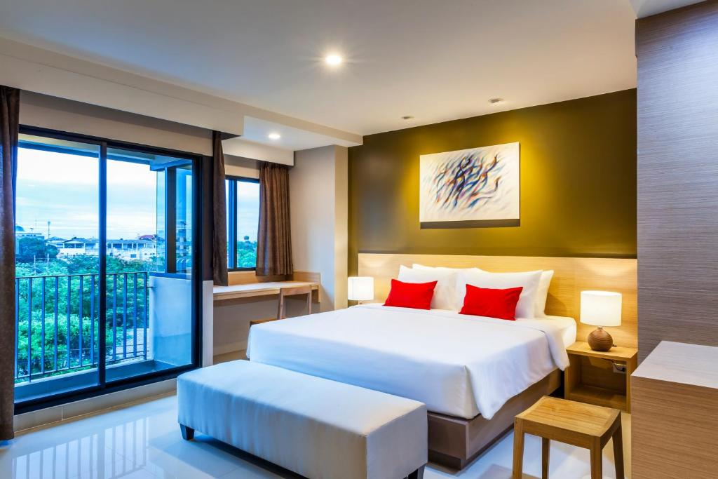 Modern hotel room with a large bed, white linens, and red accent pillows, a wooden desk by a window, bedside tables with lamps, abstract art above the bed, and a balcony overlooking greenery.