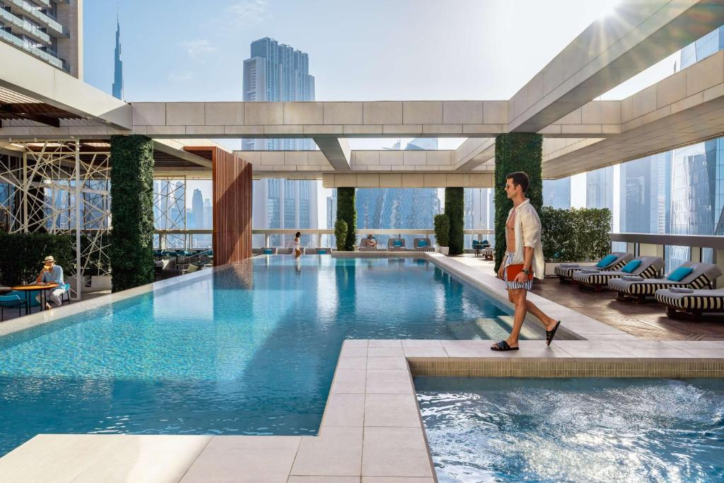 A man in casual clothes walks by a luxurious rooftop pool with lounge chairs, greenery, and distant city skyscrapers under a bright, sunny sky. Other people relax near the pool.
