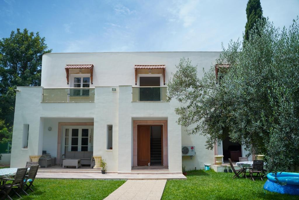 A modern two-story white house with small balconies, large windows, a wooden front door, patio furniture, and a well-kept green lawn with outdoor seating and surrounding trees.