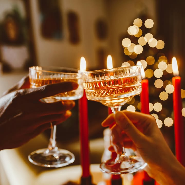 Two people clink vintage champagne glasses in a warm, festive setting. Red candles burn in the foreground, and a glowing, decorated Christmas tree is visible in the softly blurred background.