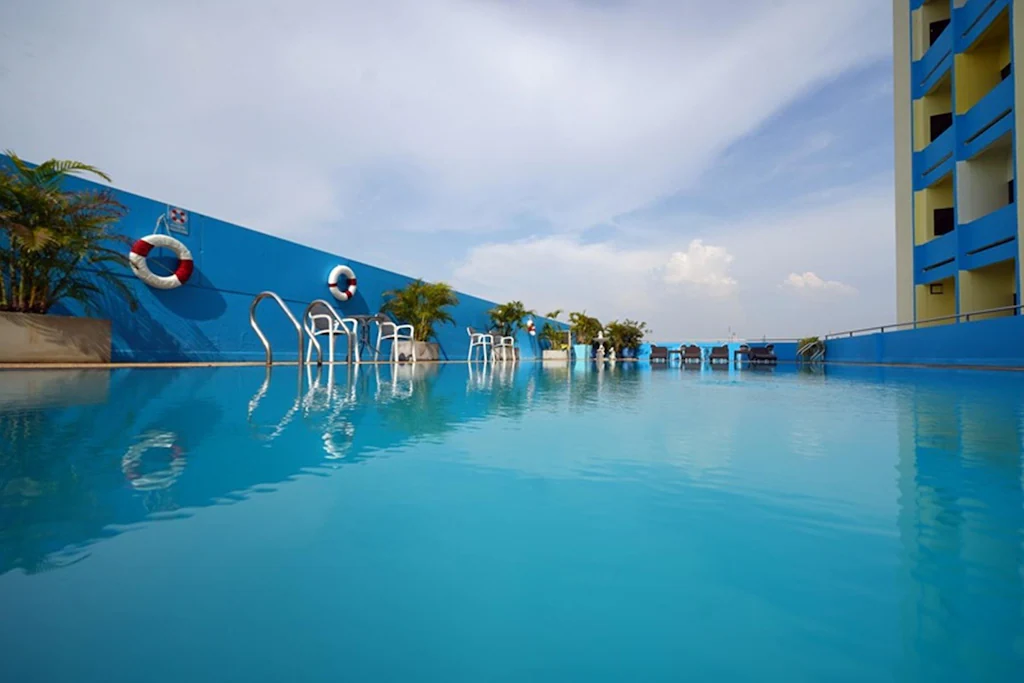 A rooftop swimming pool with clear blue water, surrounded by blue walls, safety rings, metal ladders, potted plants, and a few lounge chairs in the distance under a partly cloudy sky.