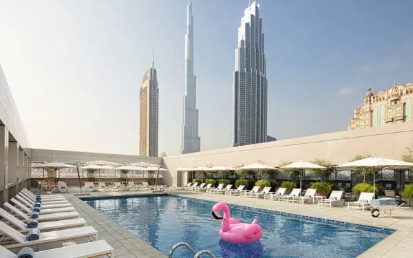 A rooftop swimming pool with white lounge chairs and umbrellas, surrounded by city skyscrapers. A large pink flamingo float is in the pool, and the Burj Khalifa is visible in the background.