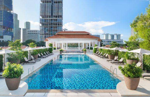 Rooftop swimming pool with blue tiles, surrounded by lounge chairs and large potted plants, set against a city skyline with tall buildings under a clear, sunny sky.