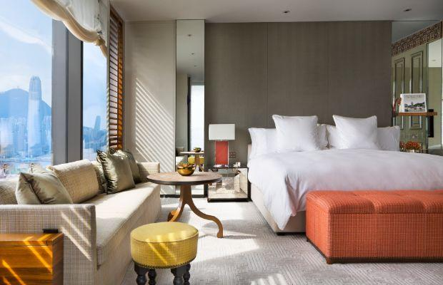 A modern hotel room with a large bed, orange bench, beige sofa, small round table, yellow stool, and floor-to-ceiling windows offering a city skyline view. The decor is neutral with warm accents and natural light fills the space.