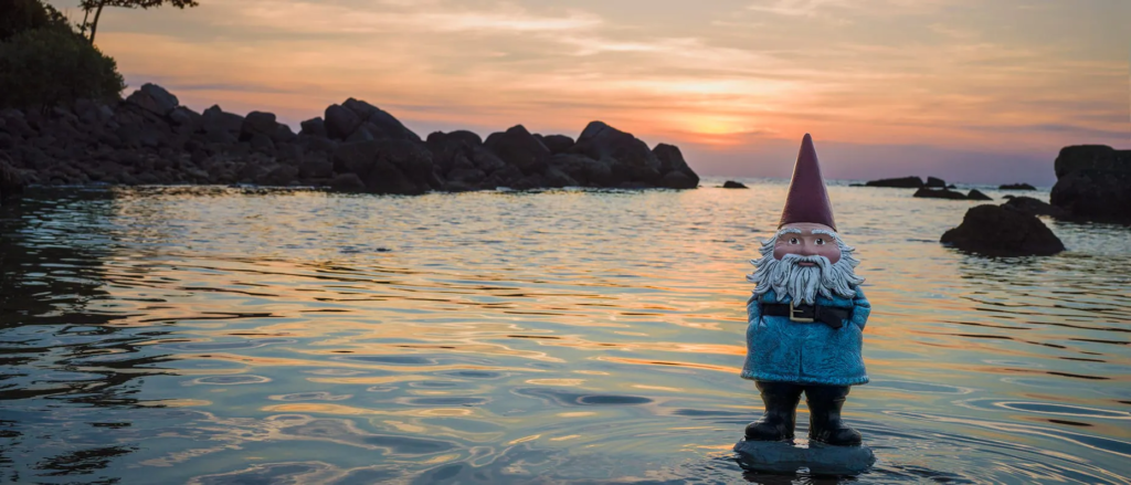 A garden gnome with a red hat and blue coat stands on a rock in shallow water at sunset, with gentle waves and dark rocks visible in the background.