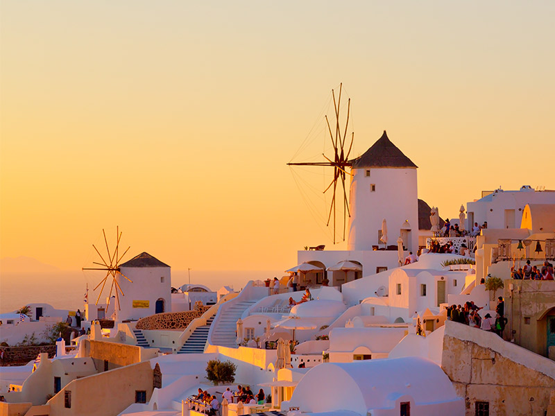 White-washed buildings and windmills on a hillside in Santorini, Greece, are bathed in warm golden light at sunset, with crowds of people enjoying the scenic view overlooking the sea.