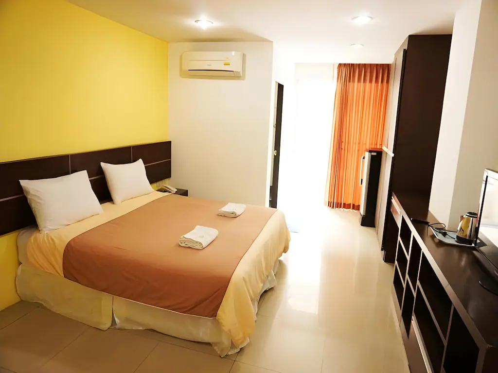 A tidy hotel room with a double bed, two white pillows, and folded towels on a beige blanket. The room has yellow and white walls, a wardrobe, TV, air conditioner, and sunlight coming through orange curtains.