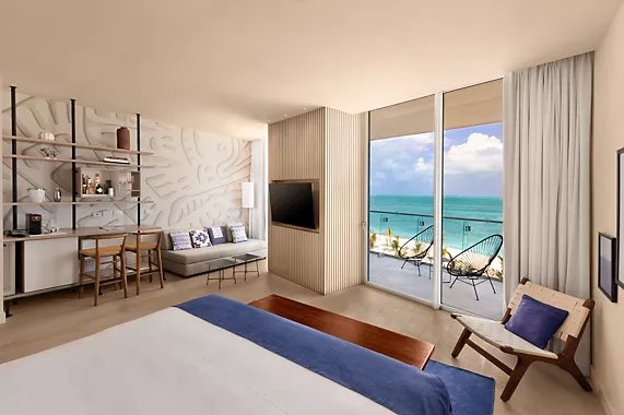 Modern hotel room with a bed, seating area, and a small table, featuring floor-to-ceiling windows that open to a balcony with ocean views. The room is decorated in neutral tones with blue accents.