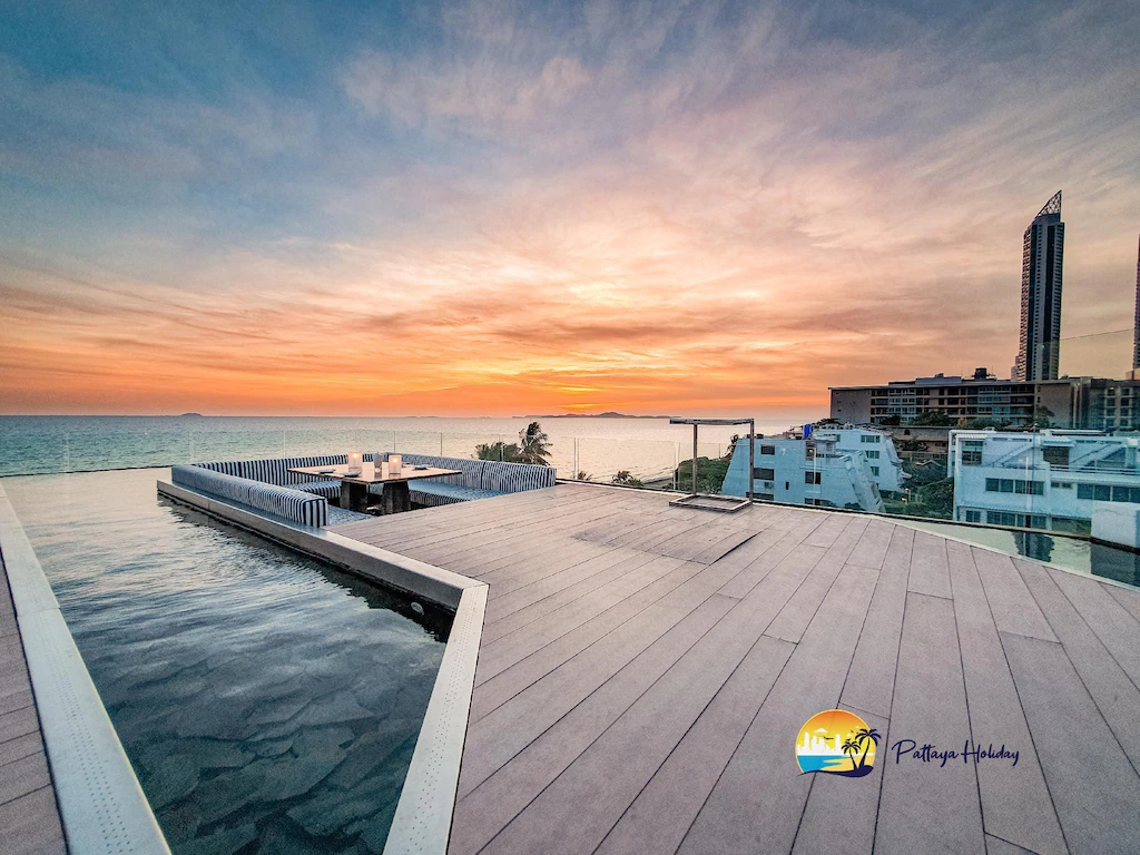 Infinity pool deck overlooking the sea at sunset, with modern buildings nearby and a high-rise in the background. The sky is colorful with orange, yellow, and blue hues. Pattaya Holiday logo is in the corner.