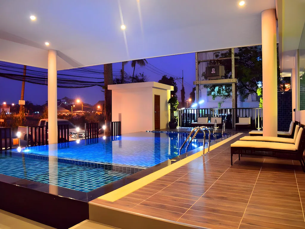 Modern indoor-outdoor pool area with blue-tiled water, poolside lounge chairs, warm lighting, and a view of city lights and trees at dusk through open walls.