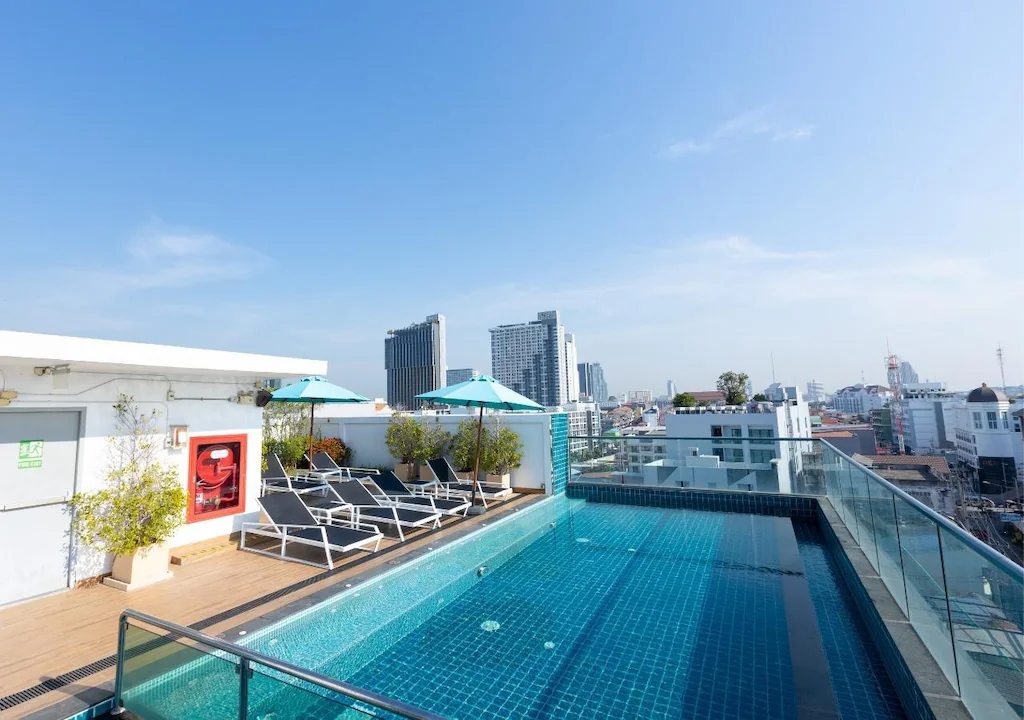 Rooftop swimming pool with clear blue water, surrounded by lounge chairs and umbrellas. City buildings and a sunny blue sky are visible in the background.