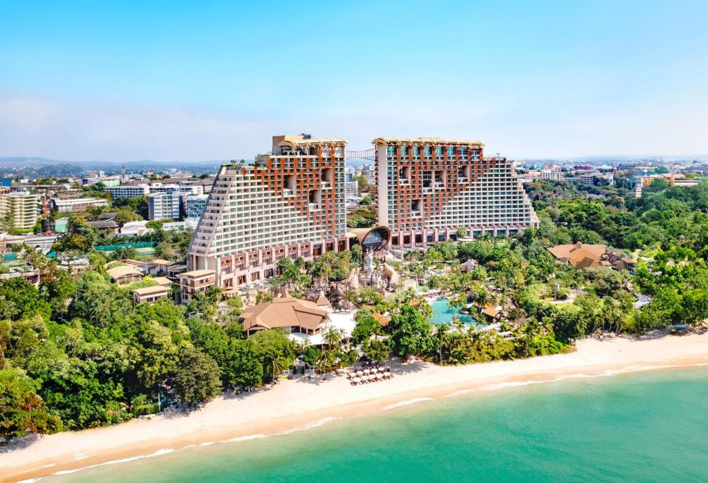 Aerial view of a large, modern beachfront resort with unique, stepped architecture, surrounded by greenery and palm trees, overlooking a sandy beach and turquoise ocean under a clear blue sky.