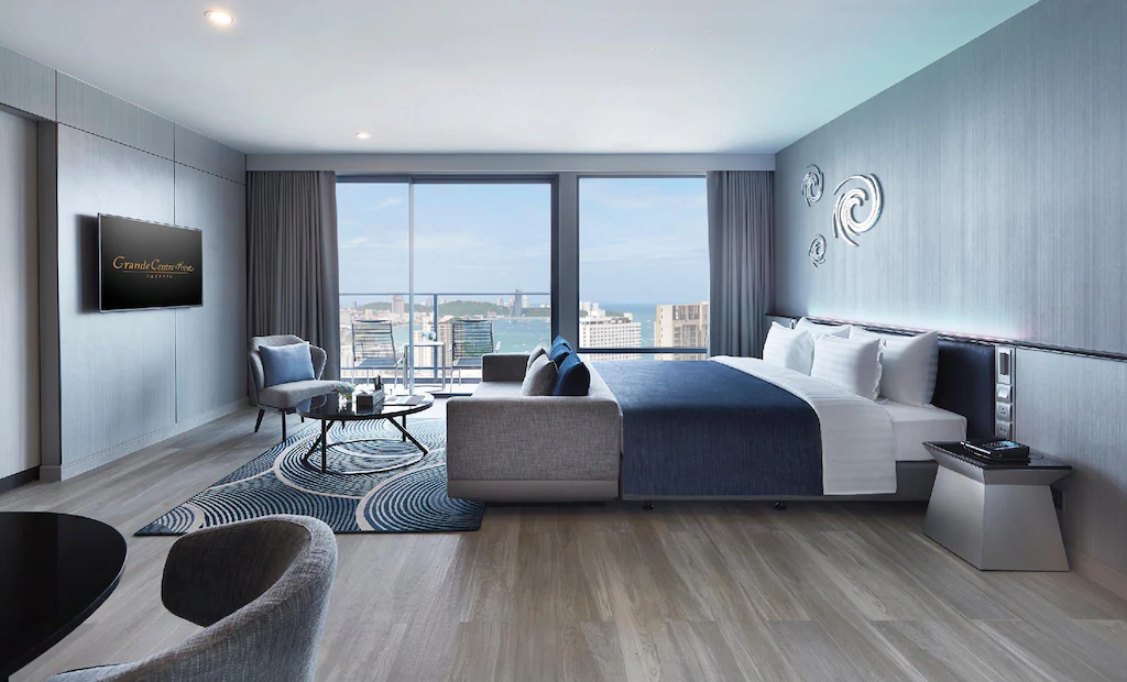 Modern hotel room with a large bed, sitting area, wall-mounted TV, and floor-to-ceiling windows offering a city and water view. Decor is neutral with blue accents and contemporary art on the wall.
