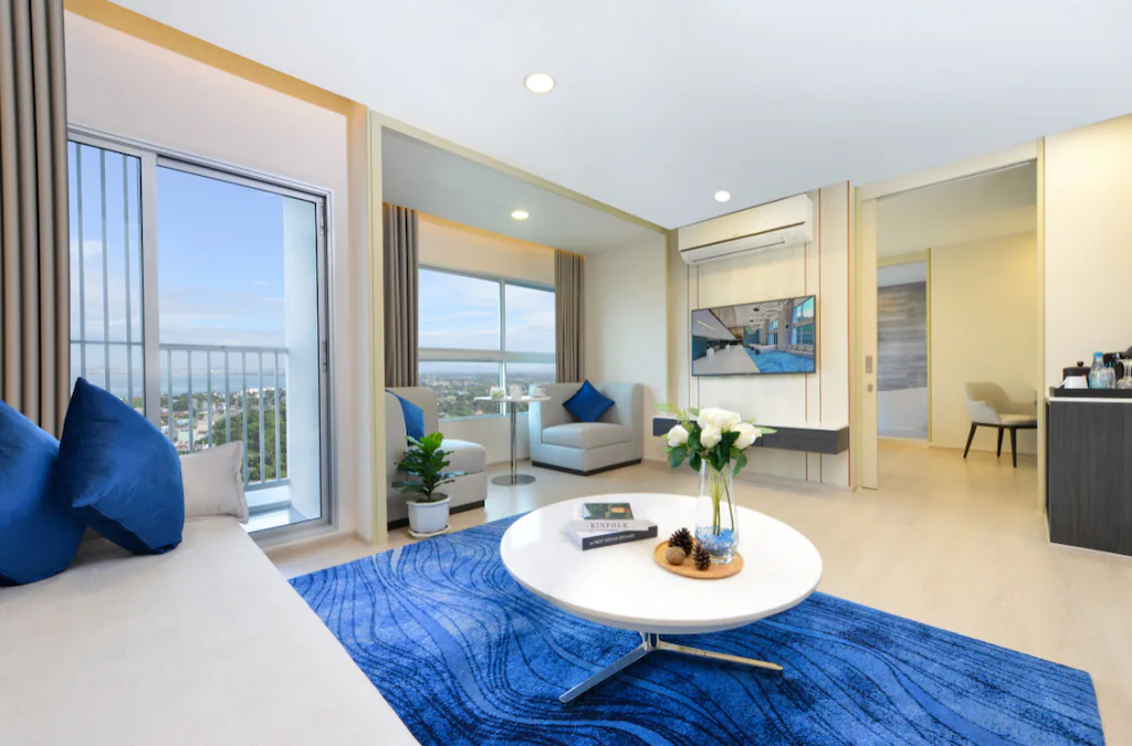 Modern, bright living room with large windows, a balcony, a white sofa with blue pillows, a blue rug, a round white coffee table with flowers, two armchairs, a wall-mounted TV, and a cityscape view.