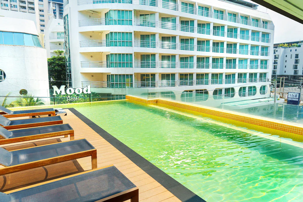 A rooftop pool with yellow tiles and clear water is surrounded by wooden lounge chairs, overlooking a modern multi-story hotel building with balconies. Mood Hotel, one of the stylish hotels in Pattaya, is visible in white letters on a wall.