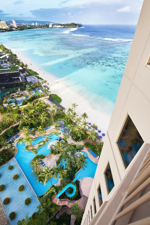 Aerial view from a tall hotel building overlooking a tropical beach, turquoise ocean, and a lush resort pool area with palm trees and winding blue pools below.