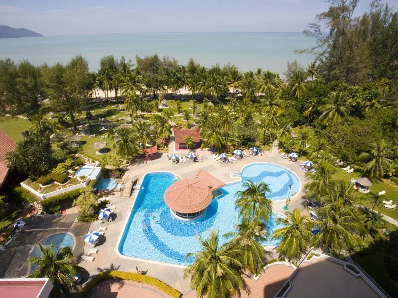 Aerial view of a tropical resort with a large outdoor pool, sun loungers, palm trees, and lush gardens near a sandy beach and calm sea under a clear sky.
