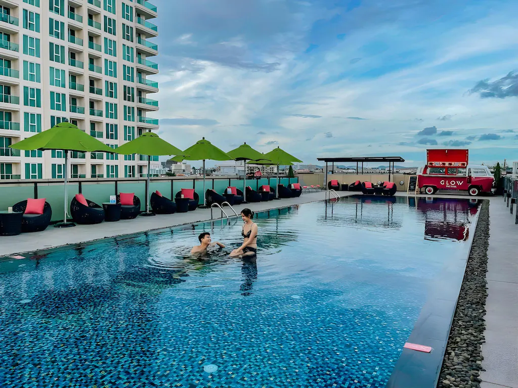 Two people swim in a rooftop pool beside a modern high-rise, enjoying green umbrellas, lounge chairs, and a red food truck—just some of the vibrant amenities found at top hotels in Pattaya under the blue, partly cloudy sky.