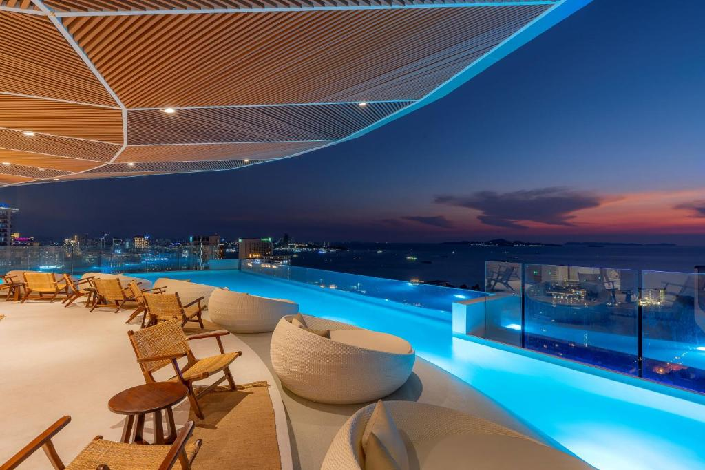 A rooftop infinity pool at one of the top hotels in Pattaya features modern lounge chairs and overlooks a vibrant city and ocean at sunset, with blue pool lights reflecting against the evening sky.