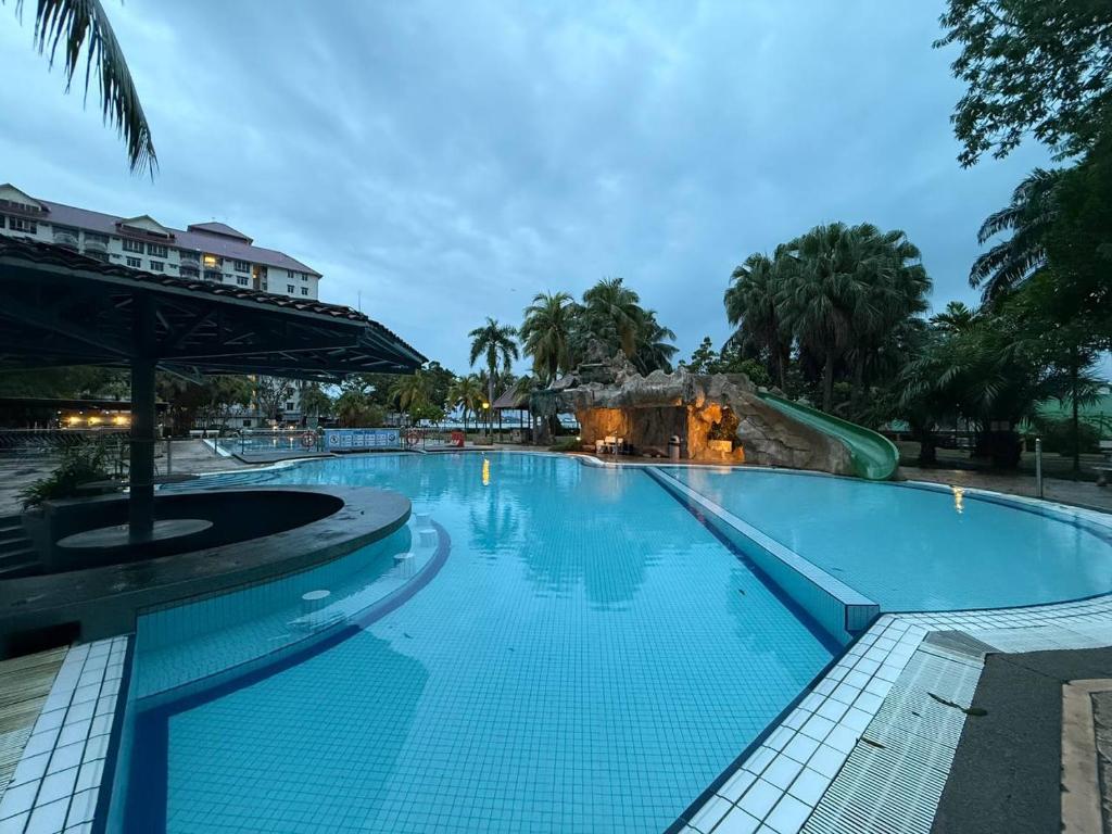 A large outdoor swimming pool with a water slide awaits at this beach resort, surrounded by palm trees and a building in the background, under a cloudy evening sky. The empty pool area is softly lit by warm lights.