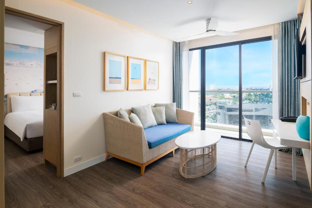 Modern hotel room with a wicker sofa, cushions, coffee table, desk, and chair. Large glass doors open to a balcony offering city and ocean views—perfect for those seeking stylish Hotels in Pattaya. A bedroom is visible through an open doorway.