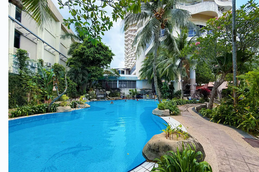Outdoor swimming pool at a beach resort, surrounded by lush tropical plants and palm trees, with a clear blue sky and tall buildings in the background. Several people are swimming and relaxing near the pool.