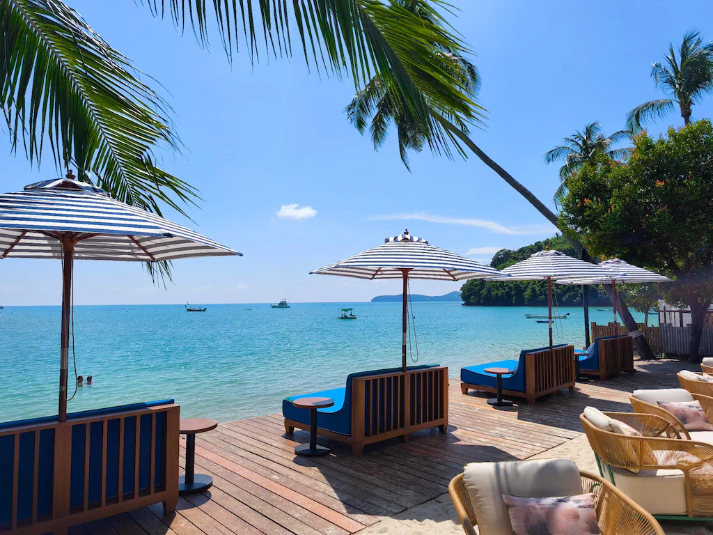 Wooden lounge chairs with striped umbrellas face a clear blue sea at a beach resort. Palm trees frame the sunny scene, and boats float in the distance under a bright blue sky.