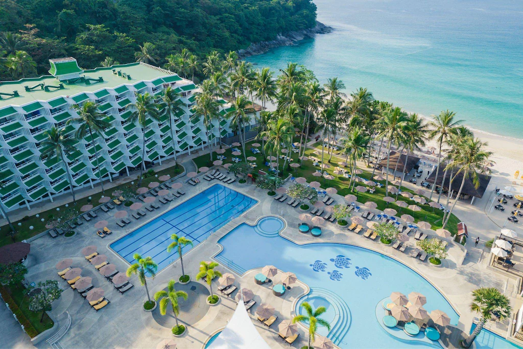 Aerial view of a beach resort with a large pool, sun loungers, palm trees, and a multistory hotel building beside a sandy beach and turquoise ocean.