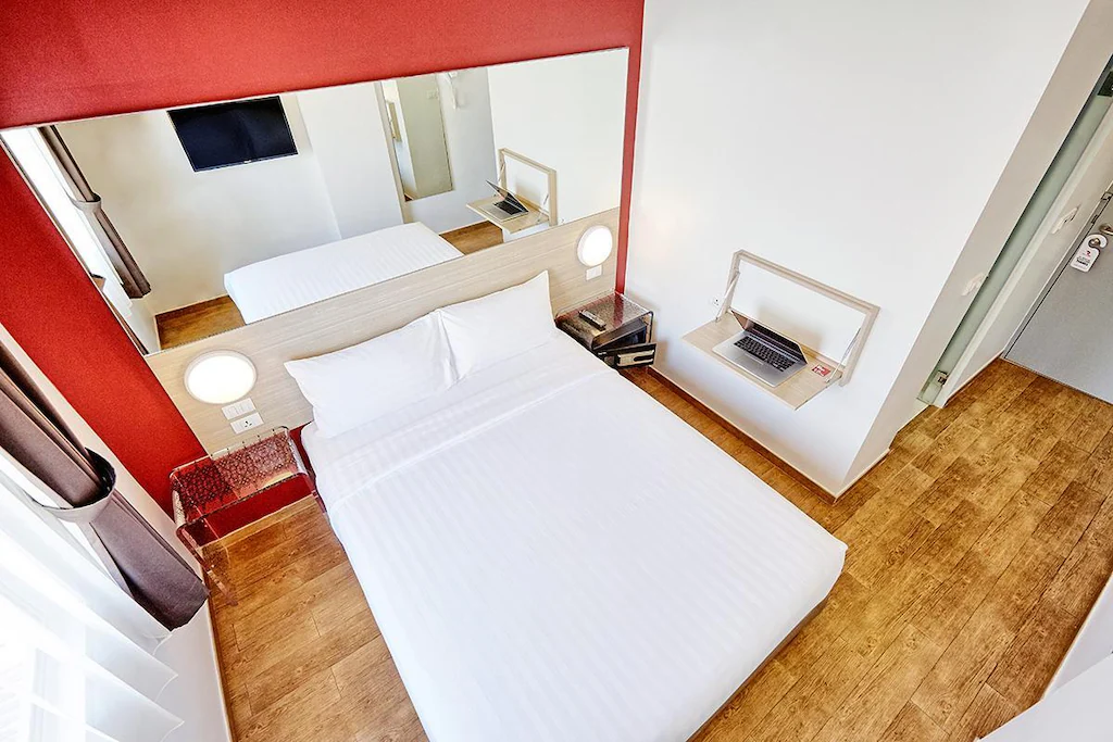 A small, modern hotel room in one of the top Hotels in Pattaya, featuring a neatly made double bed, wall-mounted TV, large mirror behind the bed, wooden floors, white walls, and a compact desk with a laptop near the door.
