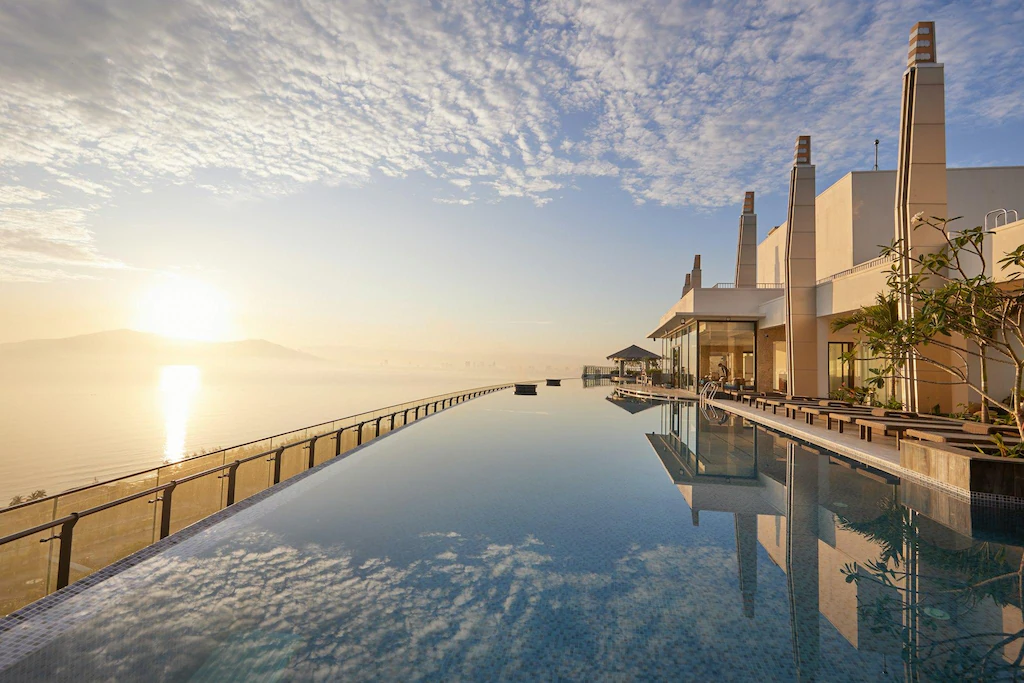 Infinity pool reflecting the sky at sunrise, with modern building and poolside lounge chairs on the right, overlooking calm water and distant mountains under a partly cloudy sky.